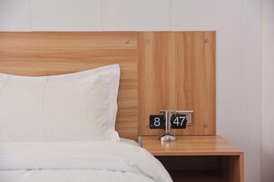 Una cama bien hecha con ropa de cama blanca y un cabecero de madera. Se ve una mesita de noche con un reloj que marca las 8:47.