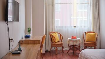 A hotel room with a wooden desk, two wicker chairs with cushions, a small table with a lace tablecloth, and a bed.
