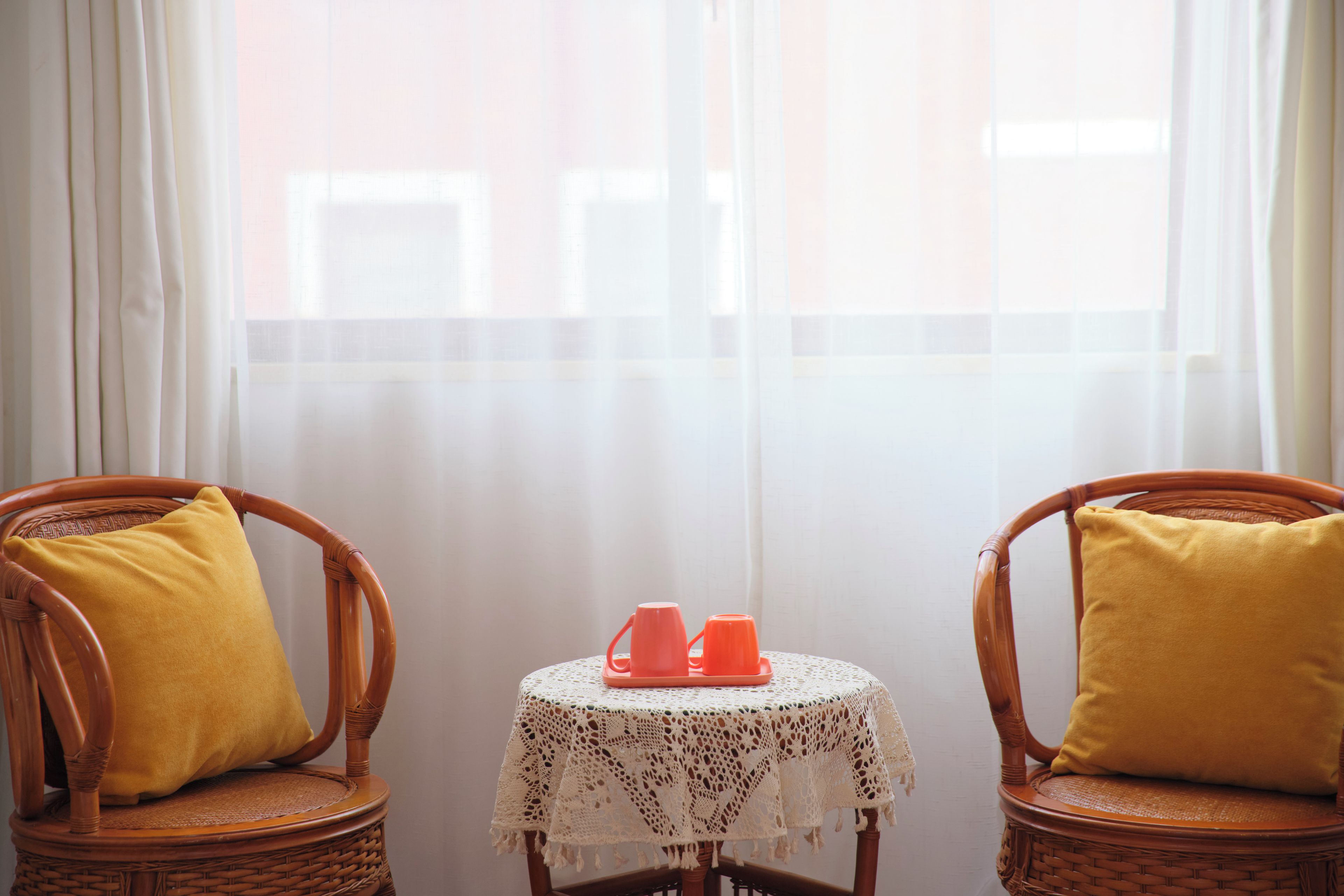 Two wicker chairs with yellow cushions, a small round table with a lace tablecloth, and two orange cups on the table.
