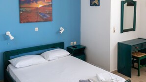 A neatly made bed with white linens, a blue wall, a painting of a sunset, and a desk with a mirror. - Aigaio View Studios & Rooms (Syros)