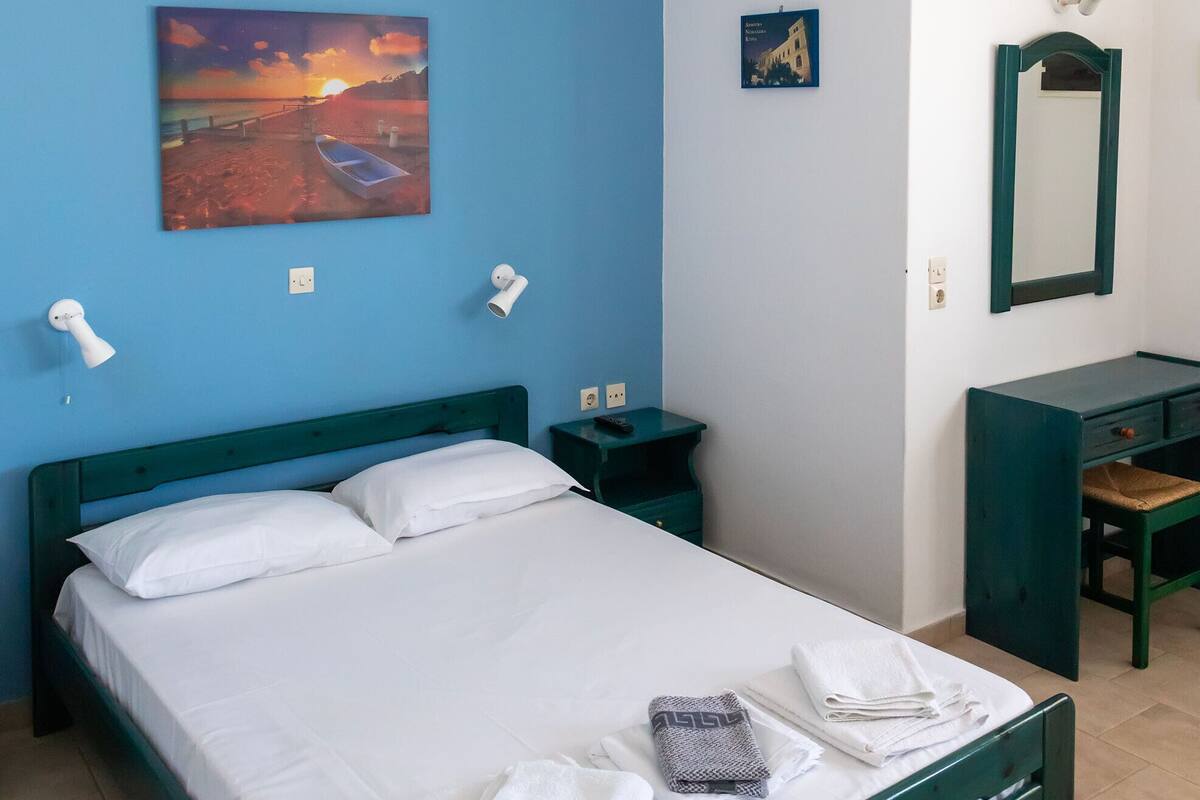 A neatly made bed with white linens, a blue wall, a painting of a sunset, and a desk with a mirror.