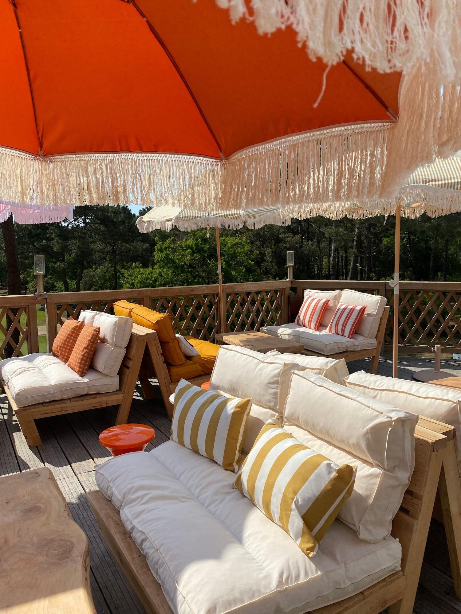 Une terrasse en bois avec des meubles d’extérieur, notamment des canapés et des fauteuils, placée sous un grand parasol à rayures.