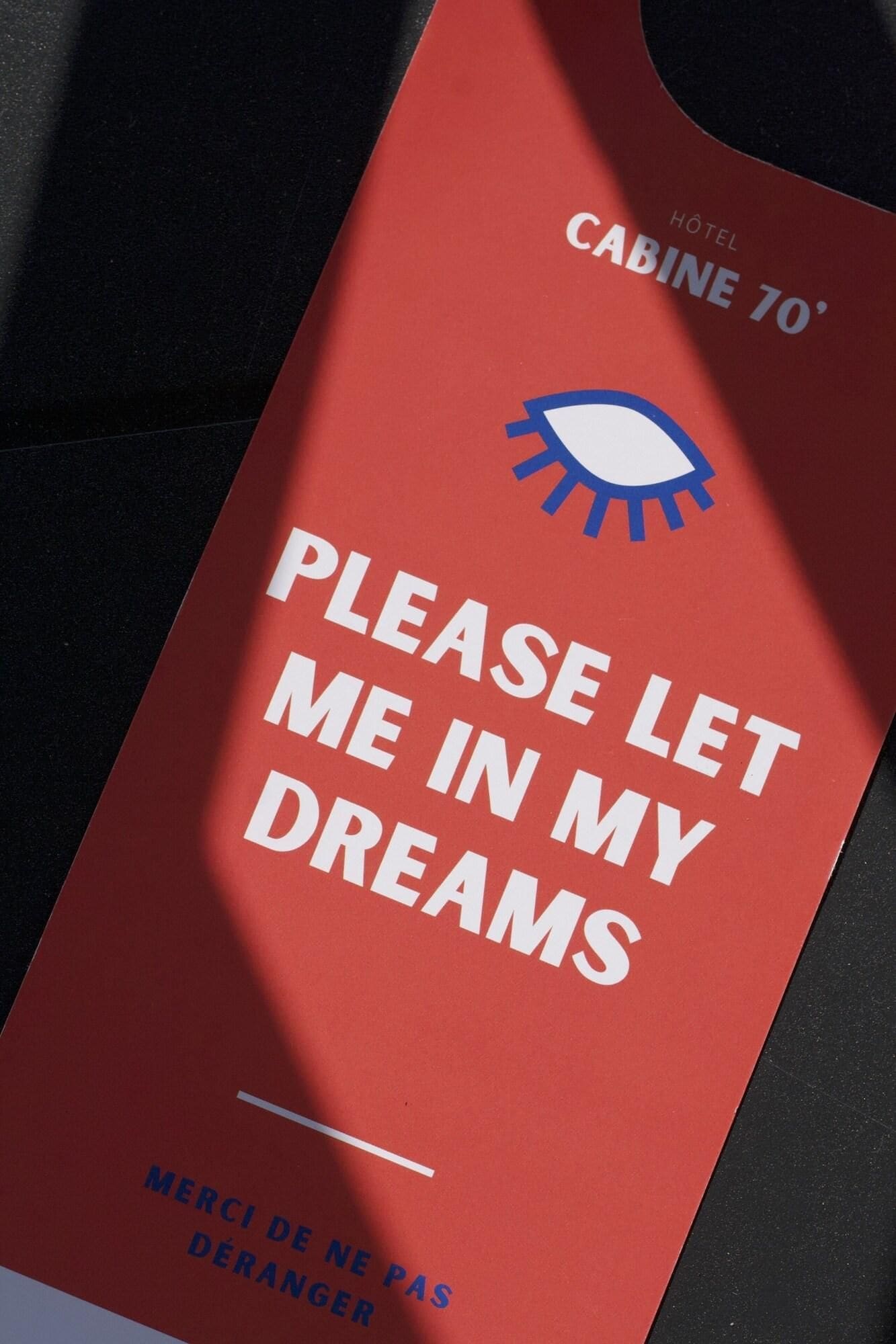 Un panneau rouge avec les inscriptions « LAISSEZ-MOI ENTRER DANS MES RÊVES » et « HÔTEL CABINE 70° ».