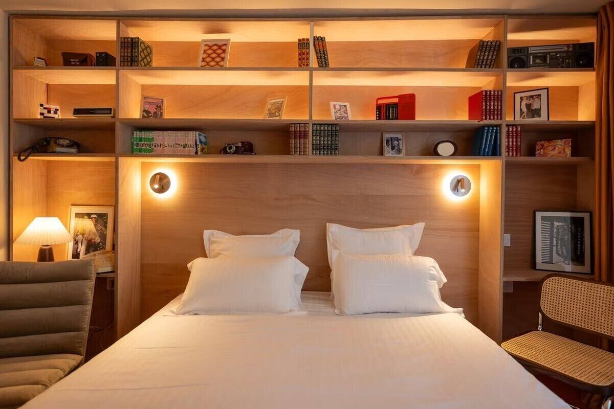 A hotel room with a large bed, two bedside tables, a wooden headboard, and a built-in bookshelf with various items.