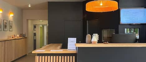 A hotel reception desk with a wooden counter, a hanging orange pendant light, and a digital screen displaying room information.