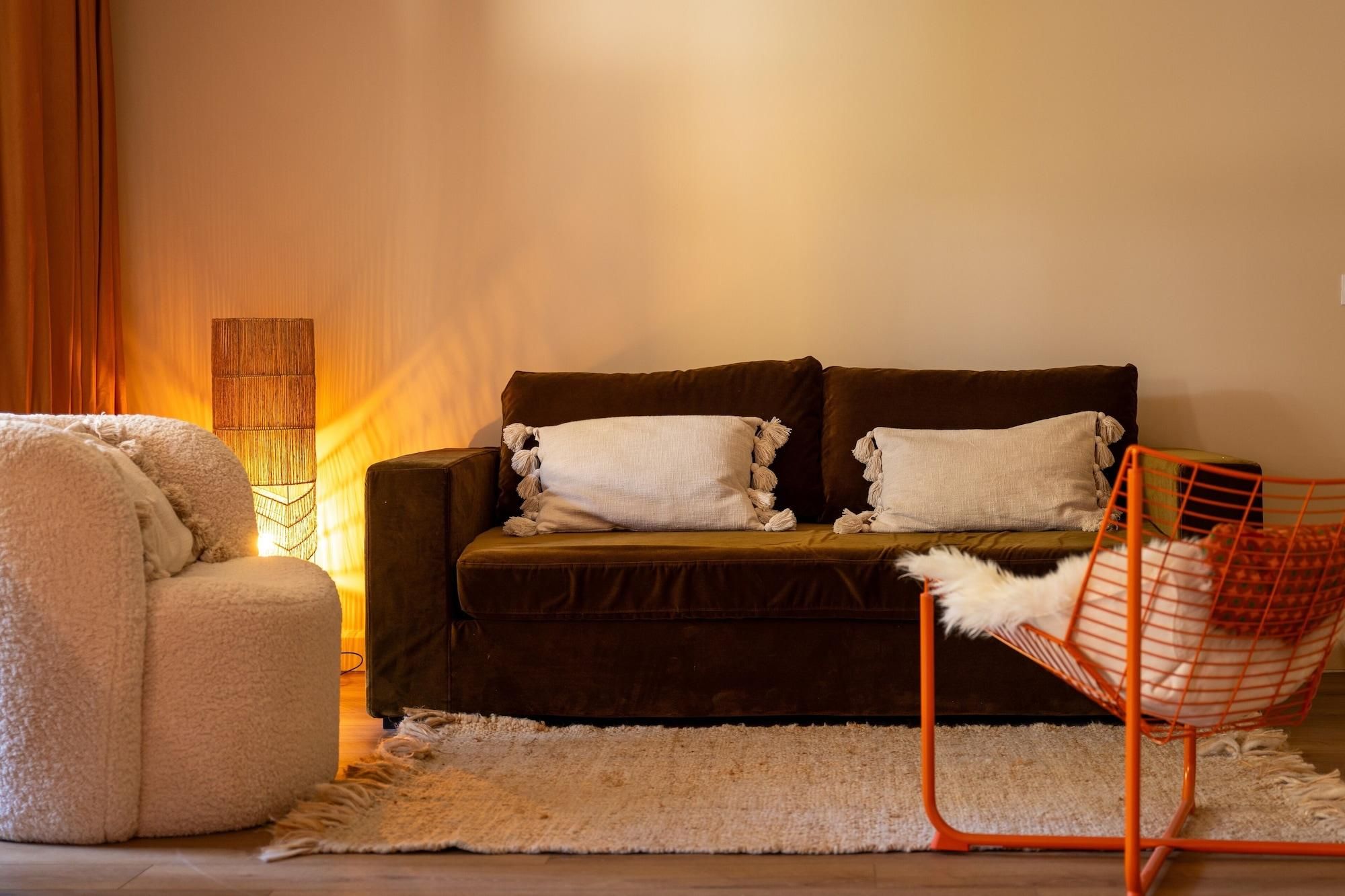 Un salon chaleureux avec un canapé marron foncé, deux coussins blancs, un fauteuil blanc moelleux et une chaise en fil de fer rouge.