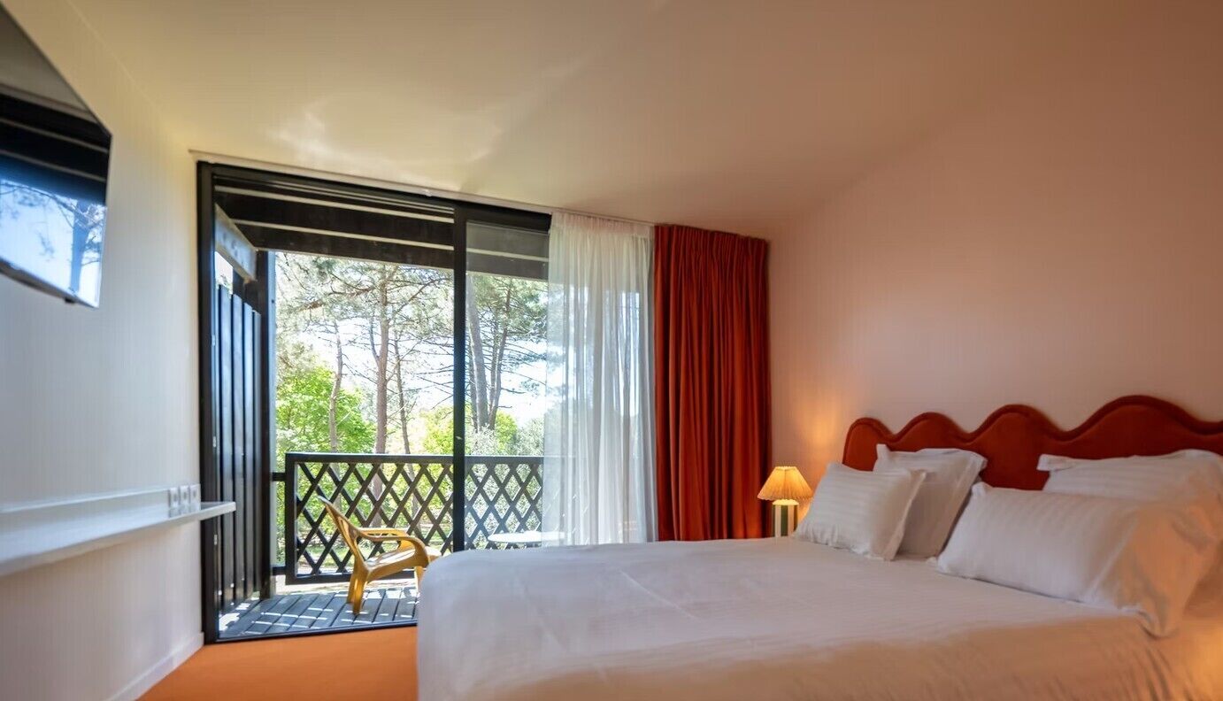 Ein Hotelzimmer mit Bett, Nachttisch, Lampe, einem Fenster mit Vorhängen und einem Balkon mit einem Stuhl.