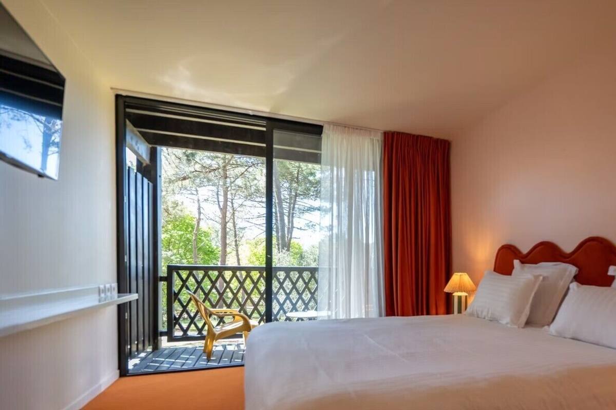 A hotel room with a bed, a nightstand, a lamp, a window with curtains, and a balcony with a chair.
