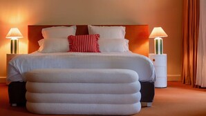 A neatly made bed with white linens and a red and white striped pillow, flanked by two bedside tables with lamps, in a warmly lit room with a large ottoman in front. - La Baignoire Hotel du Golf Lacanau (Lacanau)