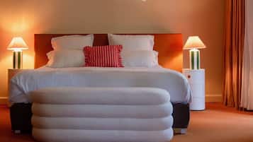 Un lit bien fait, avec du linge de lit blanc et un coussin rayé rouge et blanc, flanqué de deux tables de chevet avec des lampes, dans une pièce chaleureusement éclairée, où se trouve un grand pouf devant le lit.