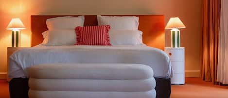 Un lit bien fait, avec du linge de lit blanc et un coussin rayé rouge et blanc, flanqué de deux tables de chevet avec des lampes, dans une pièce chaleureusement éclairée, où se trouve un grand pouf devant le lit.