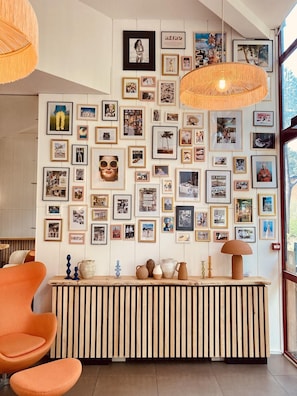 Un mur orné de nombreuses photographies encadrées, un comptoir en bois à lattes avec des objets décoratifs, et des chaises orange.