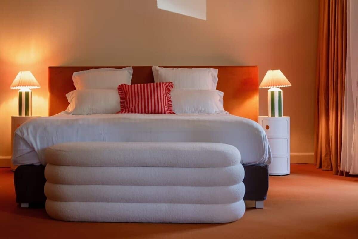 A neatly made bed with white linens and a red and white striped pillow, flanked by two bedside tables with lamps, in a warmly lit room with a large ottoman in front.