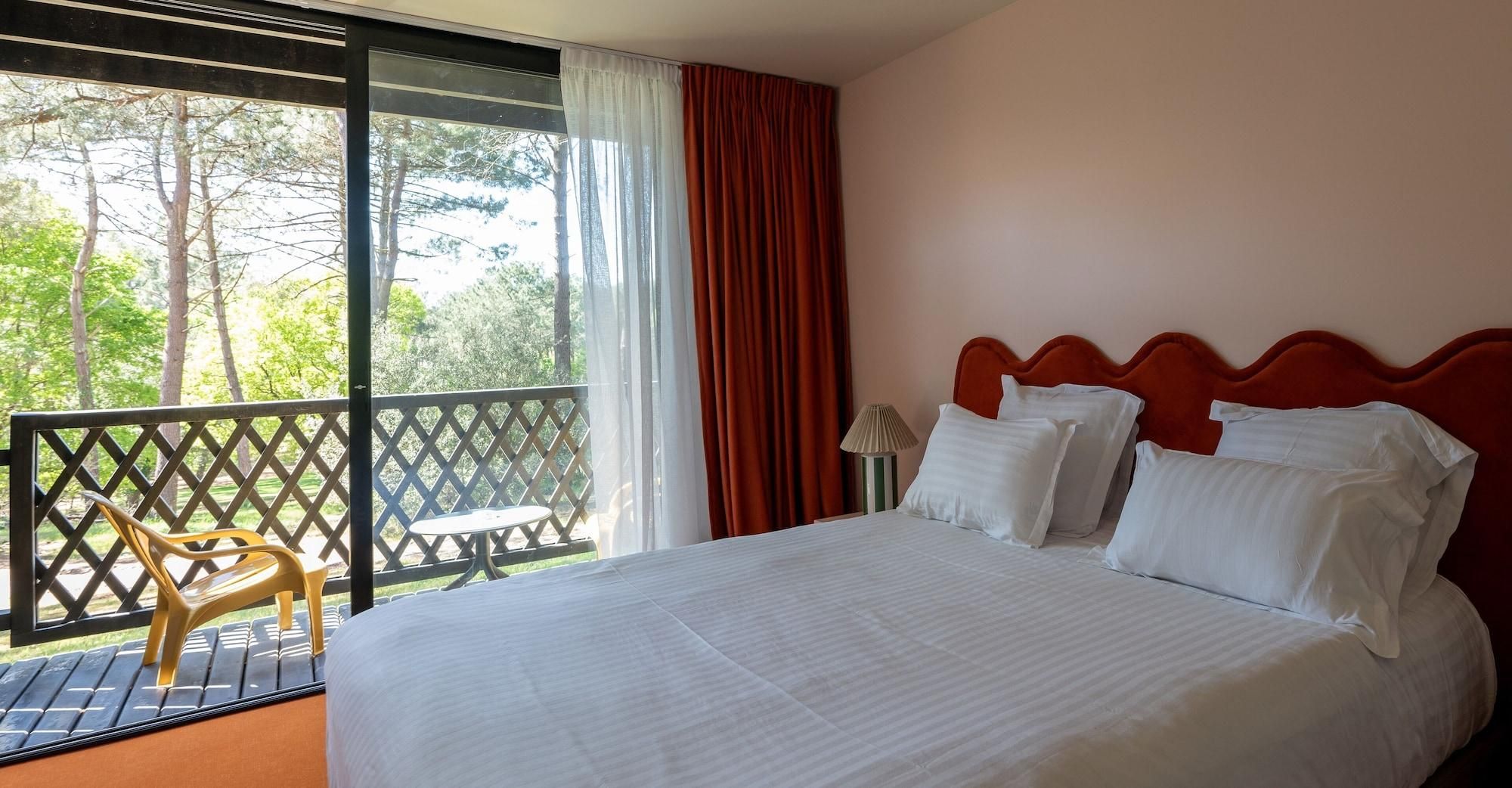 A hotel room with a bed, a view of trees, a balcony with a chair, and a table.