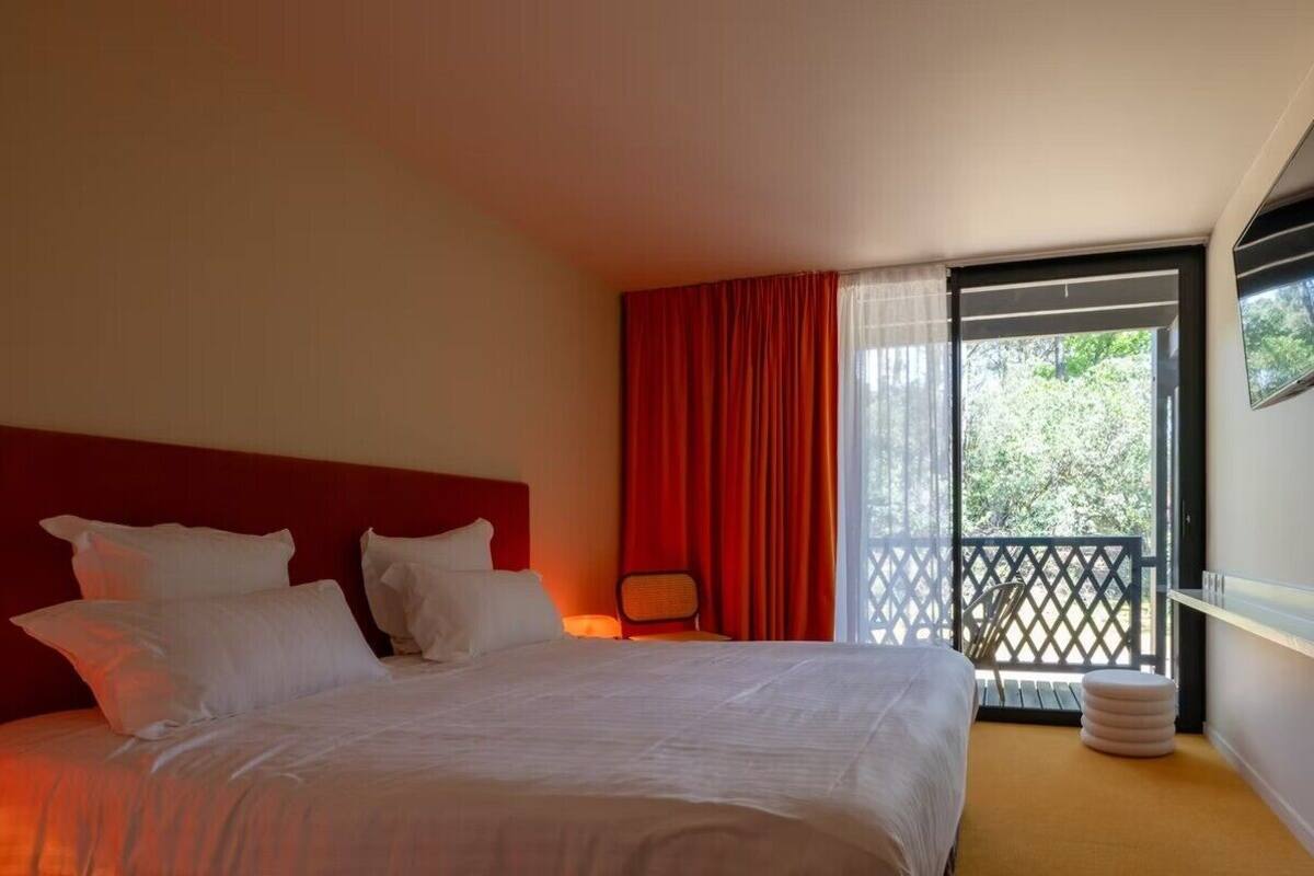 A hotel room with a large bed, a red headboard, a window with red curtains, and a balcony with a lattice railing.