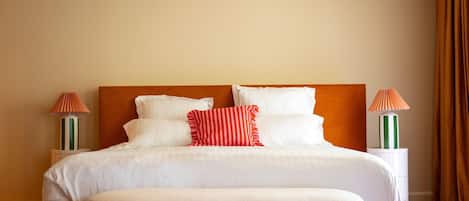 Una cama bien tendida con ropa blanca y un cojín a rayas rojas, flanqueada por dos mesitas de noche con lámparas, en una habitación con una pared lisa y una cortina marrón.