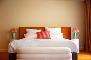 Una cama bien tendida con ropa blanca y un cojín a rayas rojas, flanqueada por dos mesitas de noche con lámparas, en una habitación con una pared lisa y una cortina marrón.