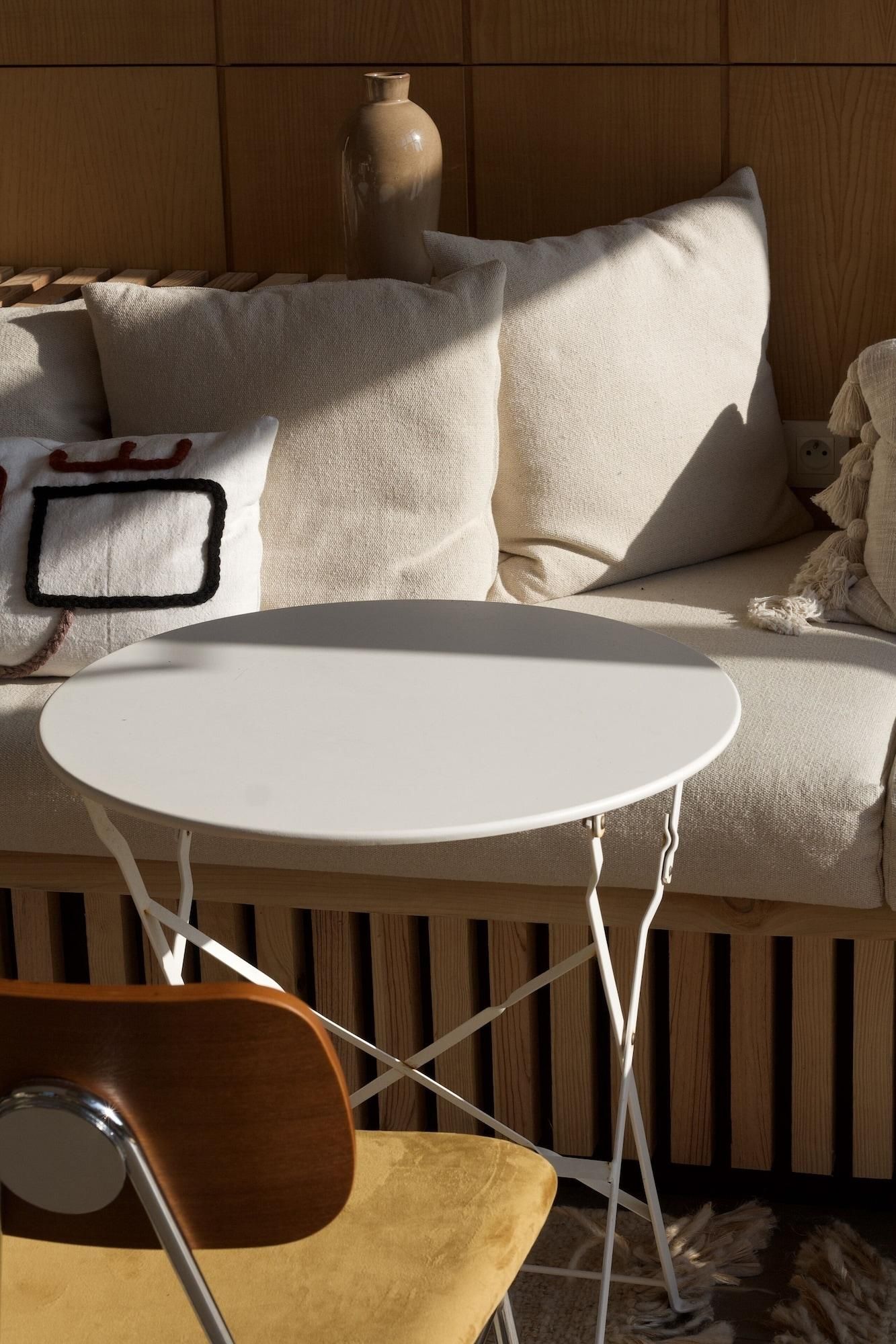 A room with a round white table, a wooden chair, a sofa with cushions, and a vase.