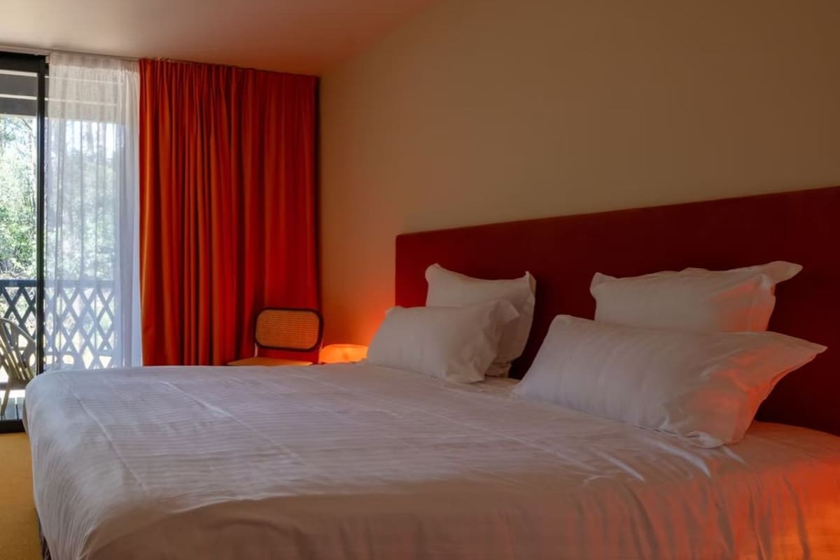 A hotel room with a large bed, red curtains, and a view of greenery outside.