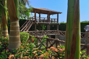 A wooden structure with a thatched roof, surrounded by lush greenery and flowering plants. - Ginger Kheda (Kheda)
