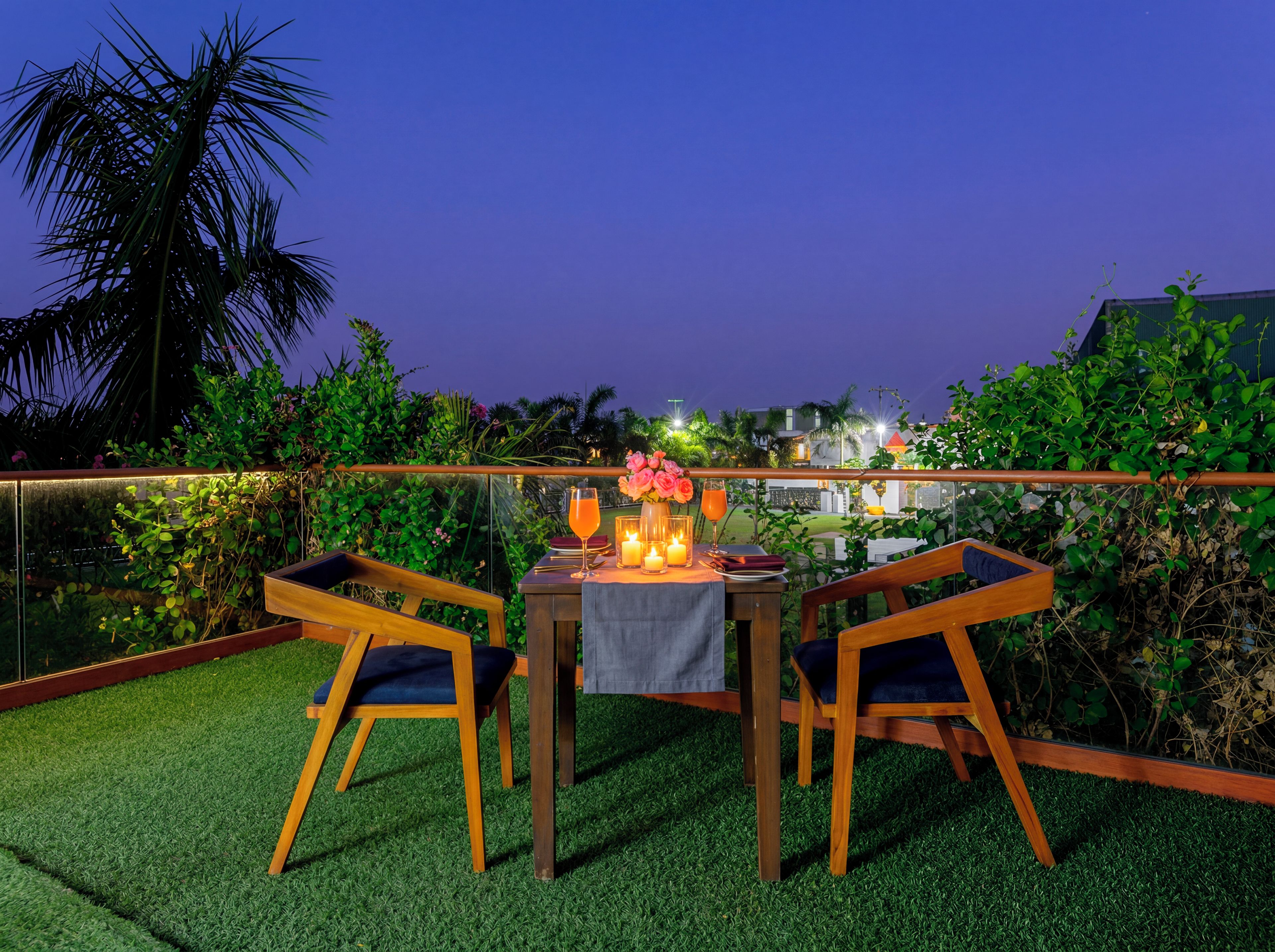 Uma área de refeições ao ar livre com mesa posta para dois, cercada por vegetação exuberante e vista para edifícios ao fundo.