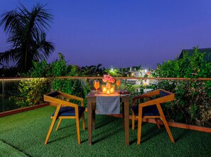 An outdoor dining area with a table set for two, surrounded by lush greenery and a view of buildings in the background. - Ginger Kheda (Kheda)