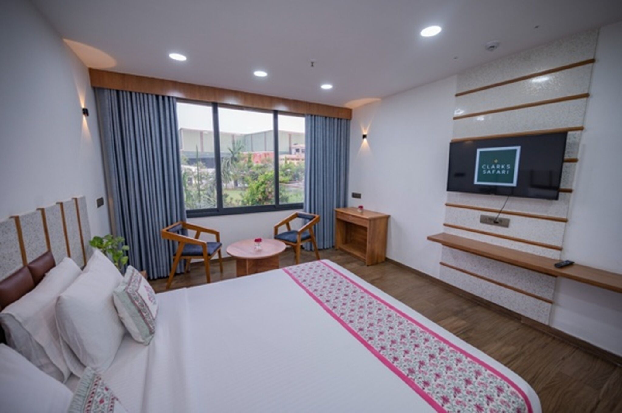 A hotel room with a large bed, a TV mounted on the wall, a small table, and a window with curtains.