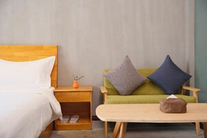 Habitación de hotel con cama de madera, un sofá verde con dos almohadas, una mesita de noche con un florero y una mesa con una caja de pañuelos.