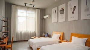 A hotel room with two beds, a desk with a chair, a small table, and a window with curtains. There are wall-mounted artworks and an air conditioning unit. - Yushan MJ Cultural Creative Theme (Huanggang)