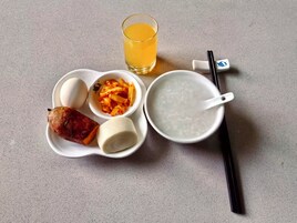 Una comida que incluye una salchicha, un plato de fideos, un vaso de jugo de naranja y un bol de arroz con palillos.