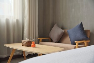 Una habitación con una mesa de madera, un sofá beige con cojines azules y marrones, y una ventana con cortinas de gasa.