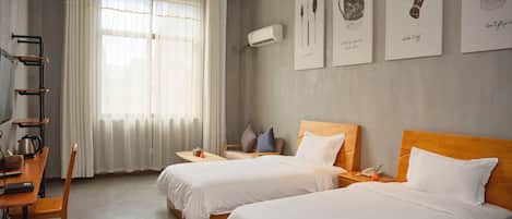 A hotel room with two beds, a desk with a chair, a small table, and a window with curtains. There are wall-mounted artworks and an air conditioning unit.