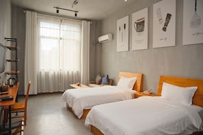 Een hotelkamer met twee bedden, een bureau met stoel, een klein tafeltje en een raam met gordijnen. Er hangen kunstwerken aan de muur en er is een airconditioning.