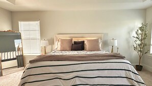 Un dormitorio con una cama grande, un ventilador de techo, una ventana con persianas, una mesita de noche, una lámpara y una planta en maceta.