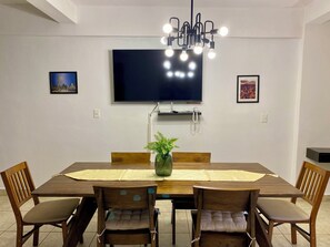 Uma área de refeições com uma mesa retangular, quatro cadeiras de madeira com assentos acolchoados, um vaso de planta e um lustre.
