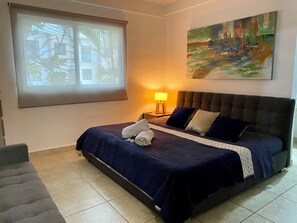 Cama bem arrumada com colcha e almofadas azul escuras, luminária de pé e um quadro na parede.