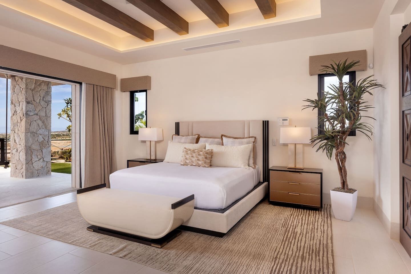 A modern bedroom with a large bed, bedside tables, a potted plant, and a view of the outdoors.