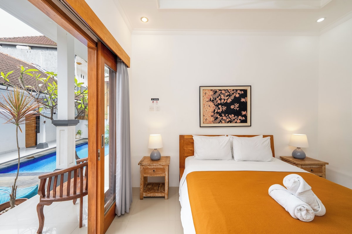 Une chambre d’hôtel avec un lit, deux tables de chevet avec des lampes, un tableau au mur et une vue sur une piscine et un espace extérieur.