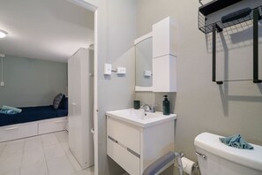Un baño pequeño con lavamanos, espejo y retrete blancos. Hay un estante pequeño sobre el lavamanos y un perchero en la pared.