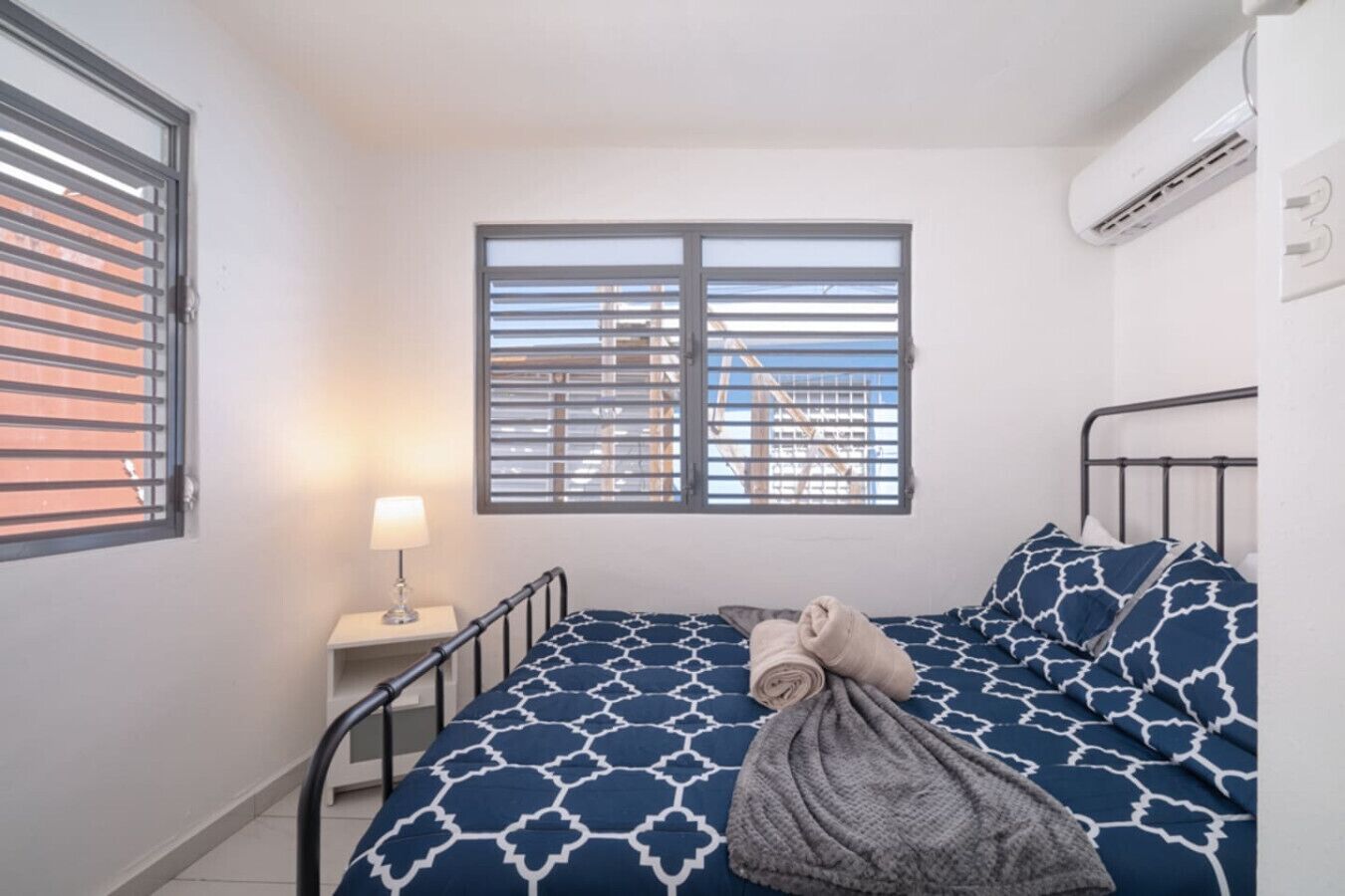 A bedroom with a bed, a window with blinds, a bedside lamp, and an air conditioning unit.