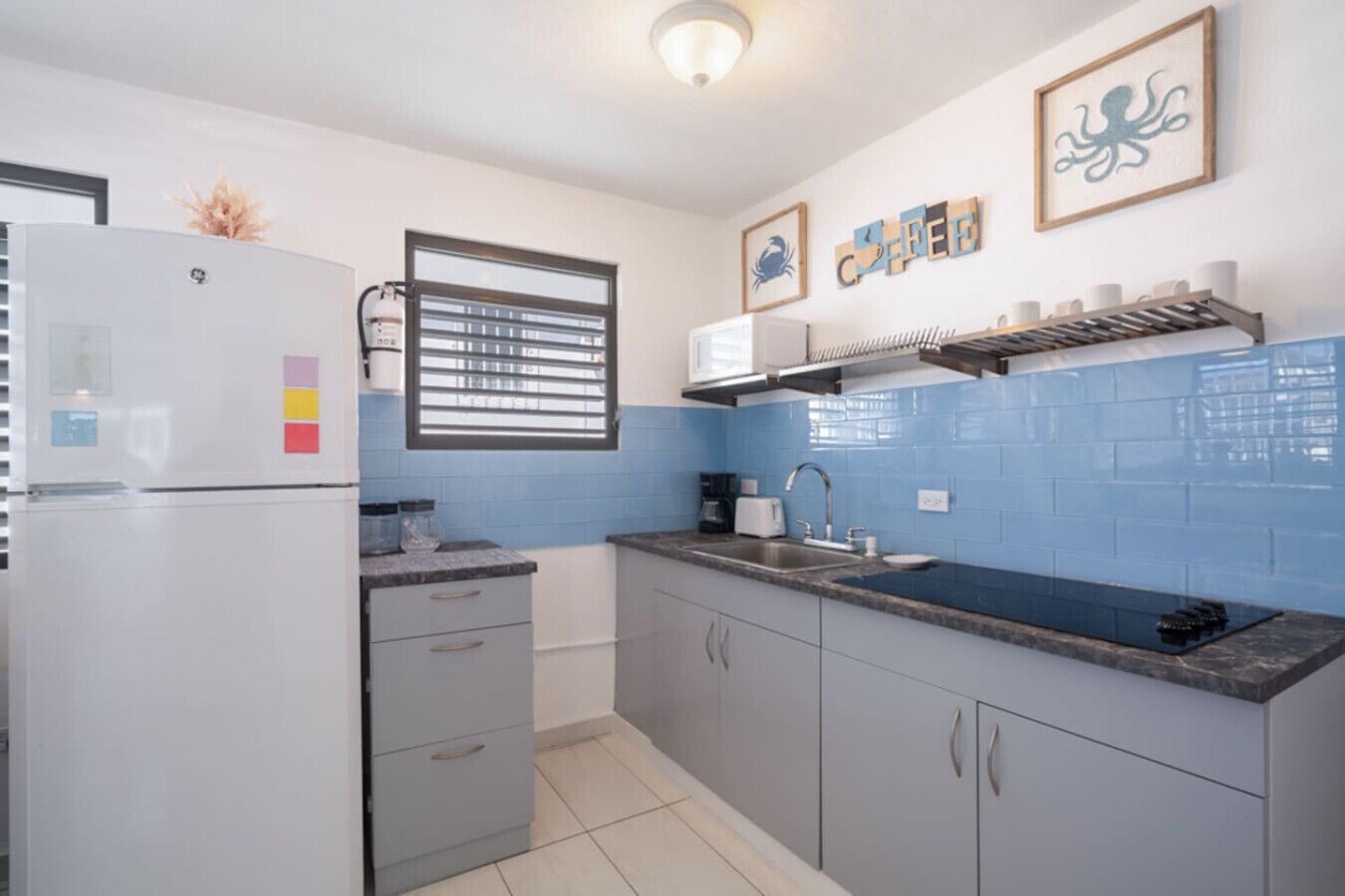 A compact kitchen with a refrigerator, a window with blinds, a microwave, a sink, and a coffee machine.