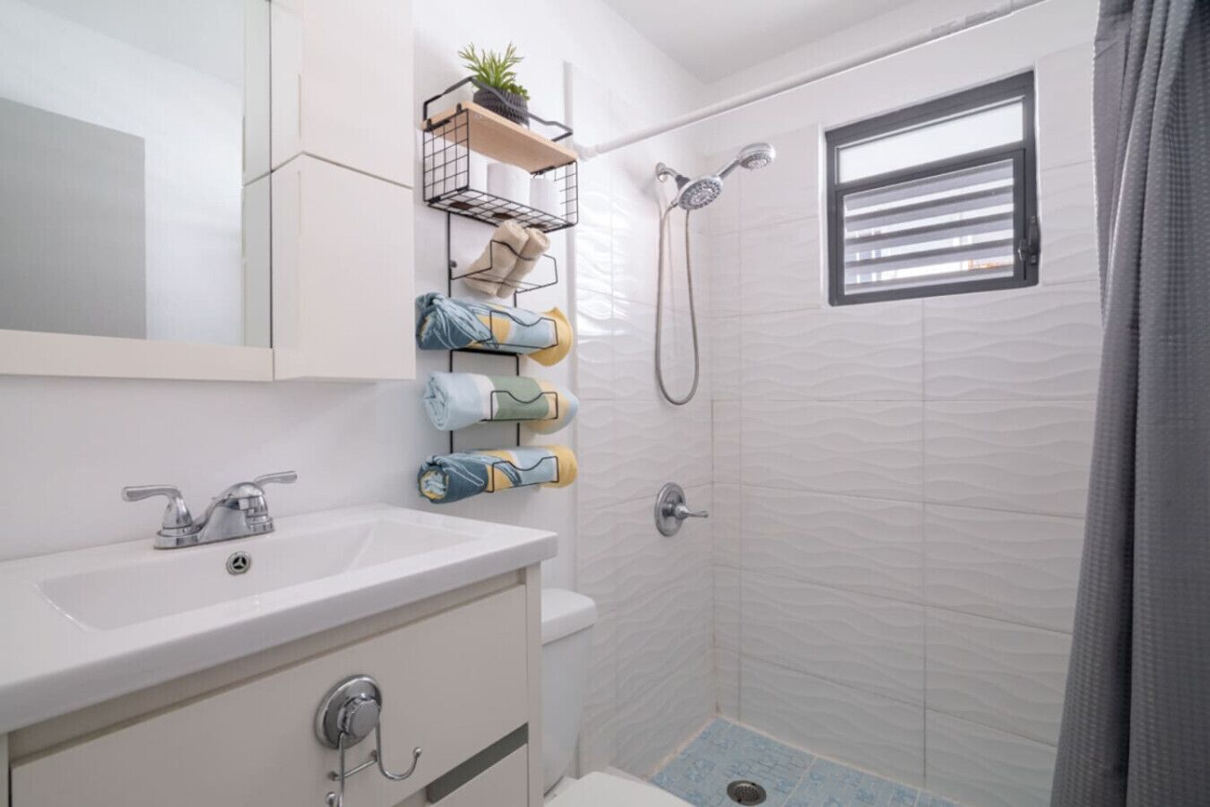 A modern bathroom with a white sink, a mirror, a shower with a handheld showerhead, and a window with blinds.