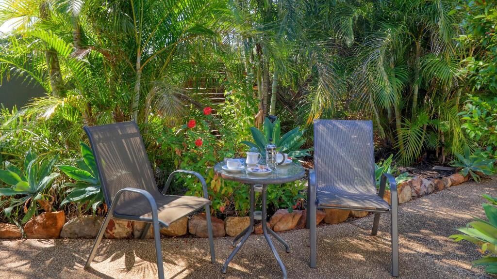 Un pequeño espacio al aire libre con dos sillas y una mesa, rodeado de exuberantes plantas tropicales y un borde de piedra.