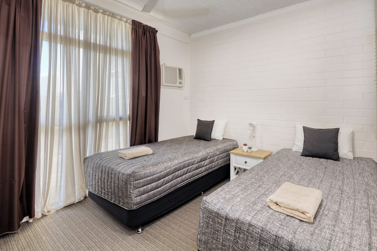 A hotel room with two beds, a window with curtains, a bedside table with a lamp, and a white brick wall.