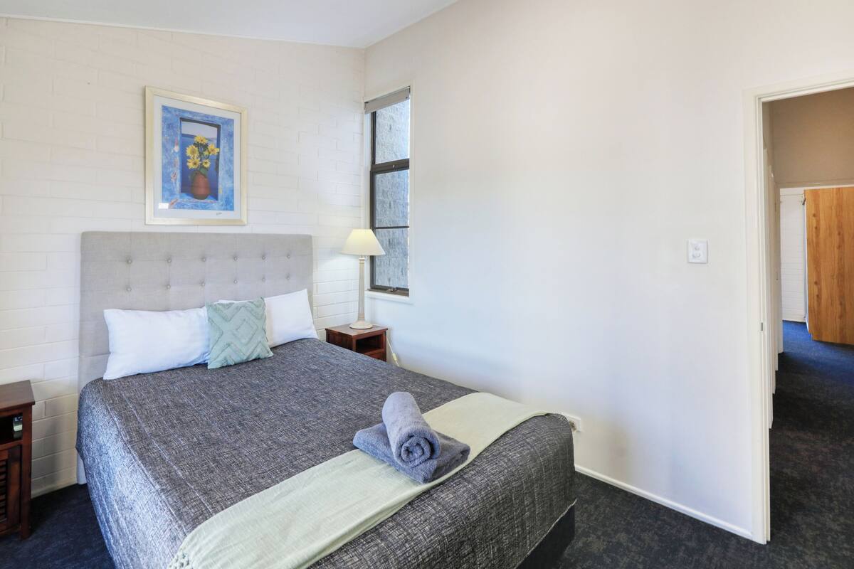 A bedroom with a bed, a nightstand, a lamp, a framed picture on the wall, and a window.