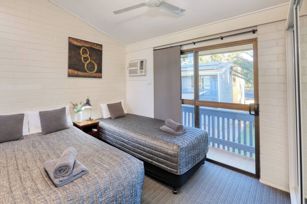 A hotel room with two beds, a ceiling fan, a window with a view of the outside, and a wall-mounted air conditioner.