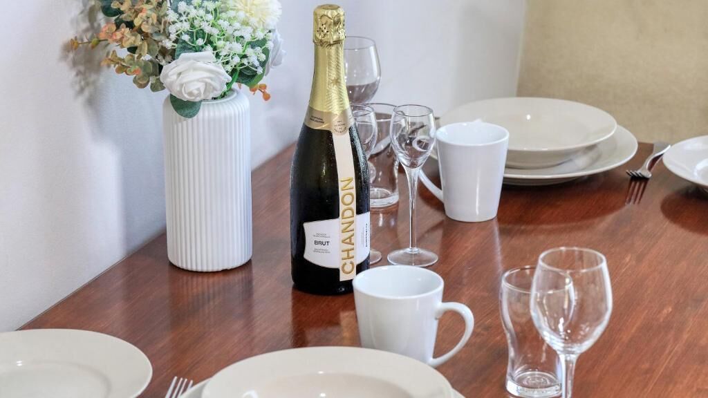 Una mesa de comedor dispuesta con platos blancos, una botella de champán, copas de vino y un jarrón con flores.