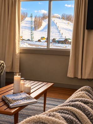 A cozy room with a large window overlooking a snowy mountain landscape, a wooden table with candles and a book, and a knitted throw on a couch. - 2BR/1BA • Fall Getaway • Across from Nub’s Nob • 10 Min to Harbor Springs (Harbor Springs)
