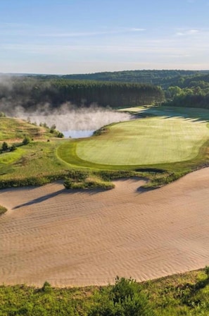 Ein Golfplatz mit Sandbunkern, einem Wasserhindernis und bewaldeten Hügeln im Hintergrund.