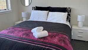 Ein ordentlich gemachtes Bett mit einer schwarz-pinkfarbenen Tagesdecke, zwei weißen Kissen und zwei zusammengerollten Handtüchern. Ein Nachttisch mit einer Lampe und einer kleinen weißen Schublade steht daneben. Ein runder Spiegel befindet sich an der Wand über dem Bett.